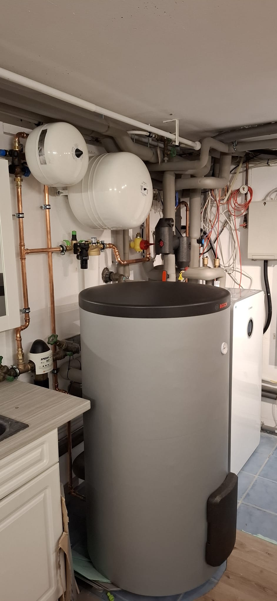 Bosch-Pufferspeicher mit Heizungseinheit, Rohren und Wandarmaturen in Technikraum mit gefliestem Boden.